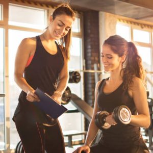trainer Female fitness instructor showing exercise progress on clipboard to young athletic woman at gym.
